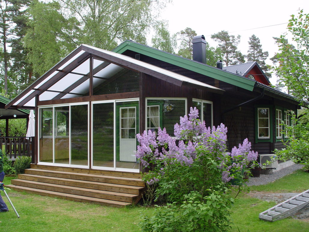 Uterum i trä Uterum med skjubara glasdörrar på ett brunt hus omgivet av en grön gräsmattas och blommande, lila, syrener.